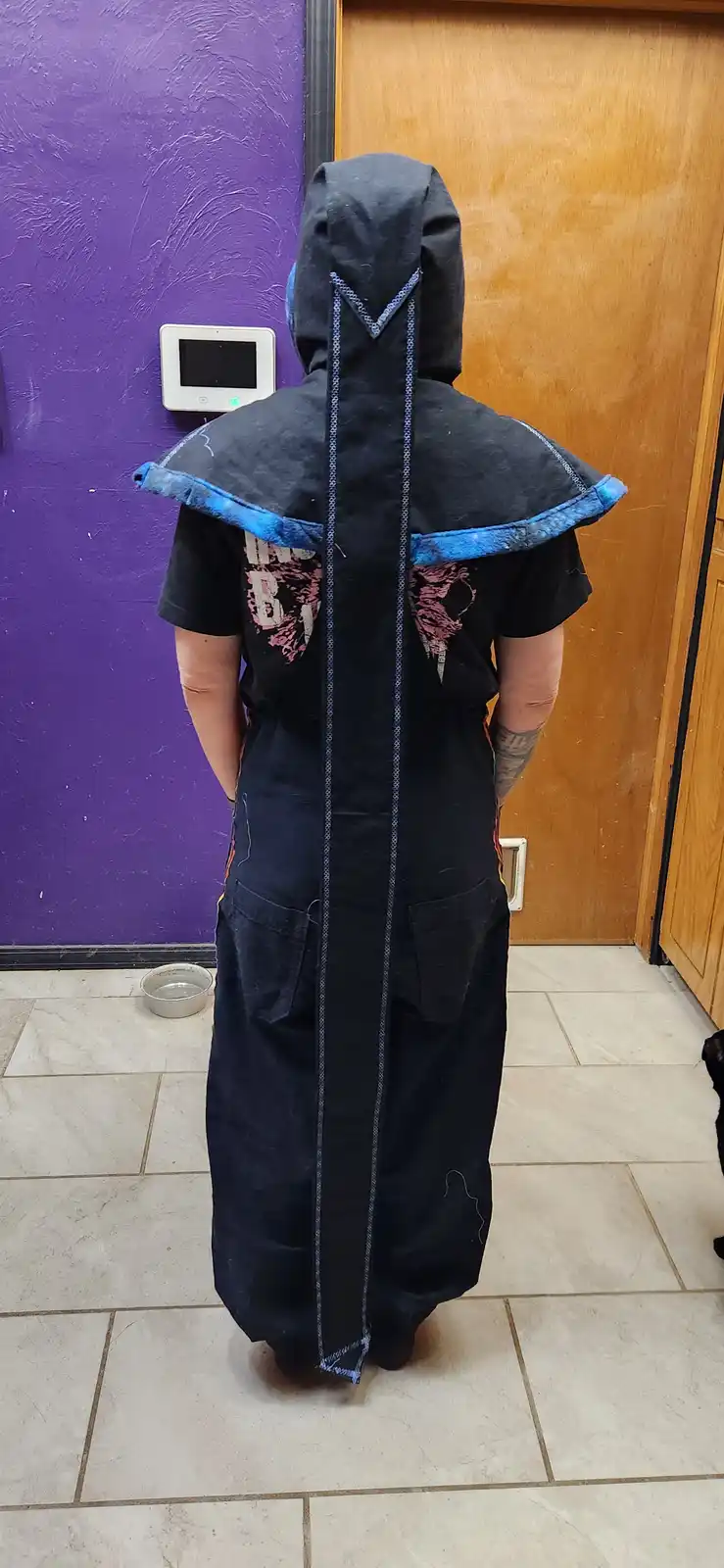 A person stands with their back to the camera, wearing a dark blue robe made from a hoodie with blue trim, revealing the…