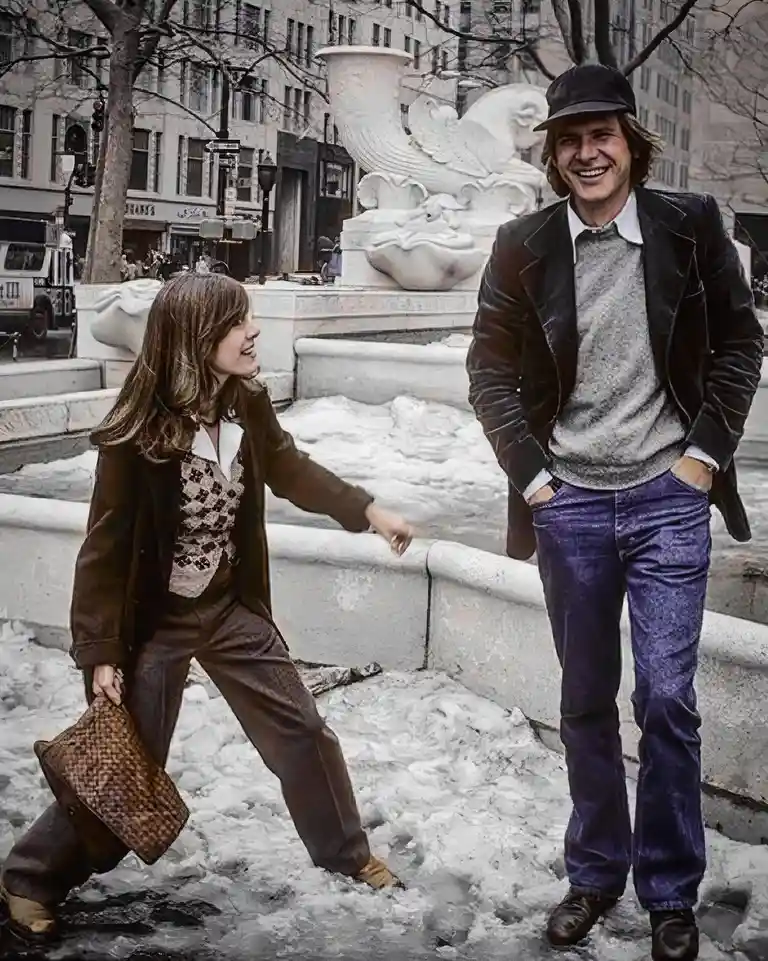 A vintage photo shows a woman in a brown suit and patterned vest stepping into icy water while holding a woven bag, smiling…