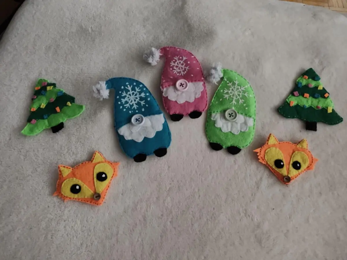 Several handmade felt ornaments lie on a white textured surface: two green Christmas trees with colorful decorations, three…