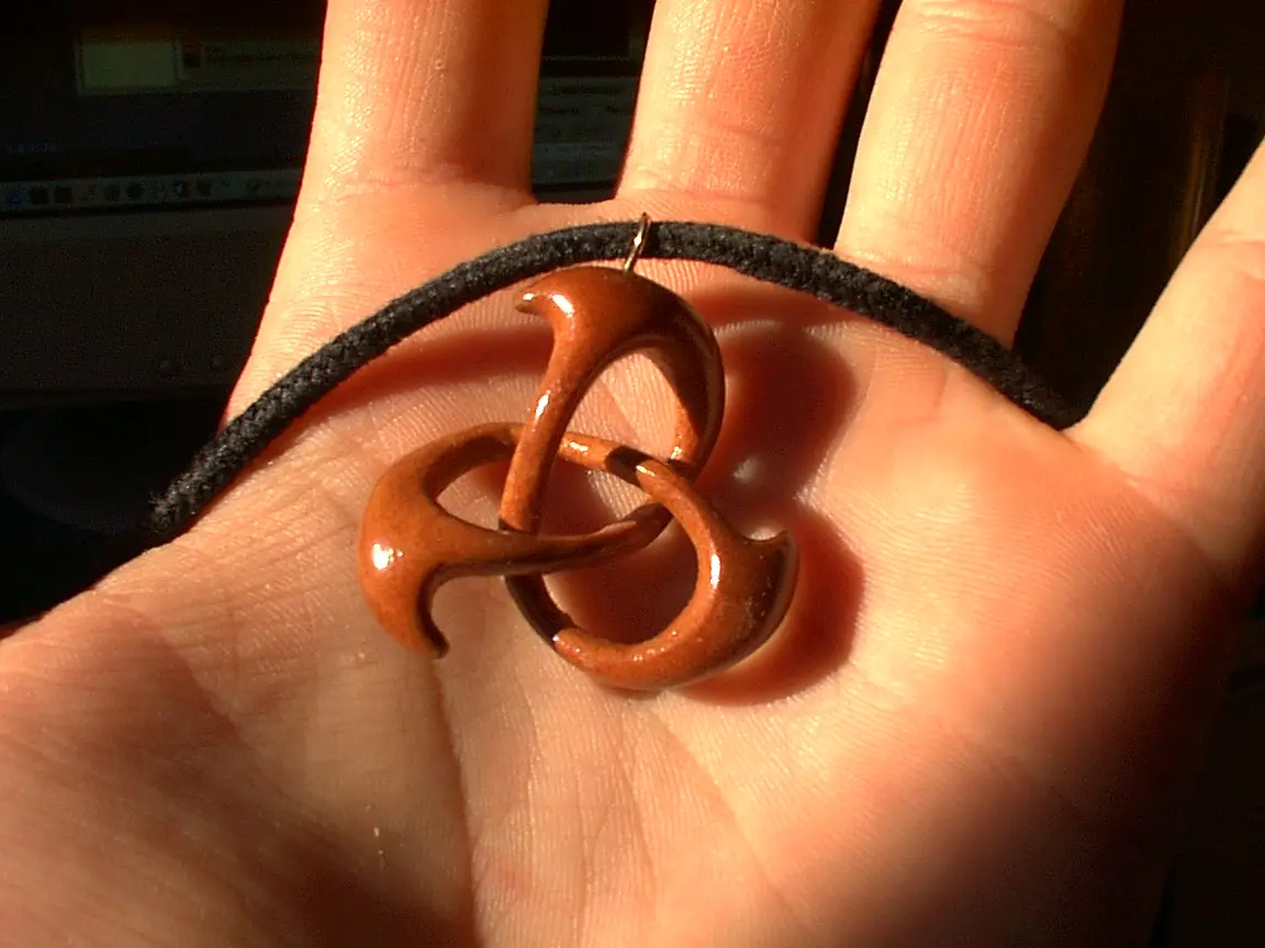 A close-up photo shows a hand holding a brown, glossy, three-looped knot-shaped pendant on a black cord. The pendant's…