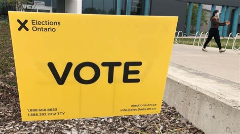 A large yellow Elections Ontario sign with the word 'VOTE' in bold black letters stands outdoors on a sidewalk. In the…
