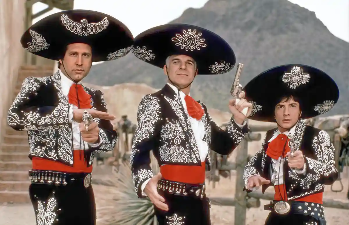 three men dressed in ornate black mariachi outfits with large sombreros and red sashes stand in a desert setting, each…