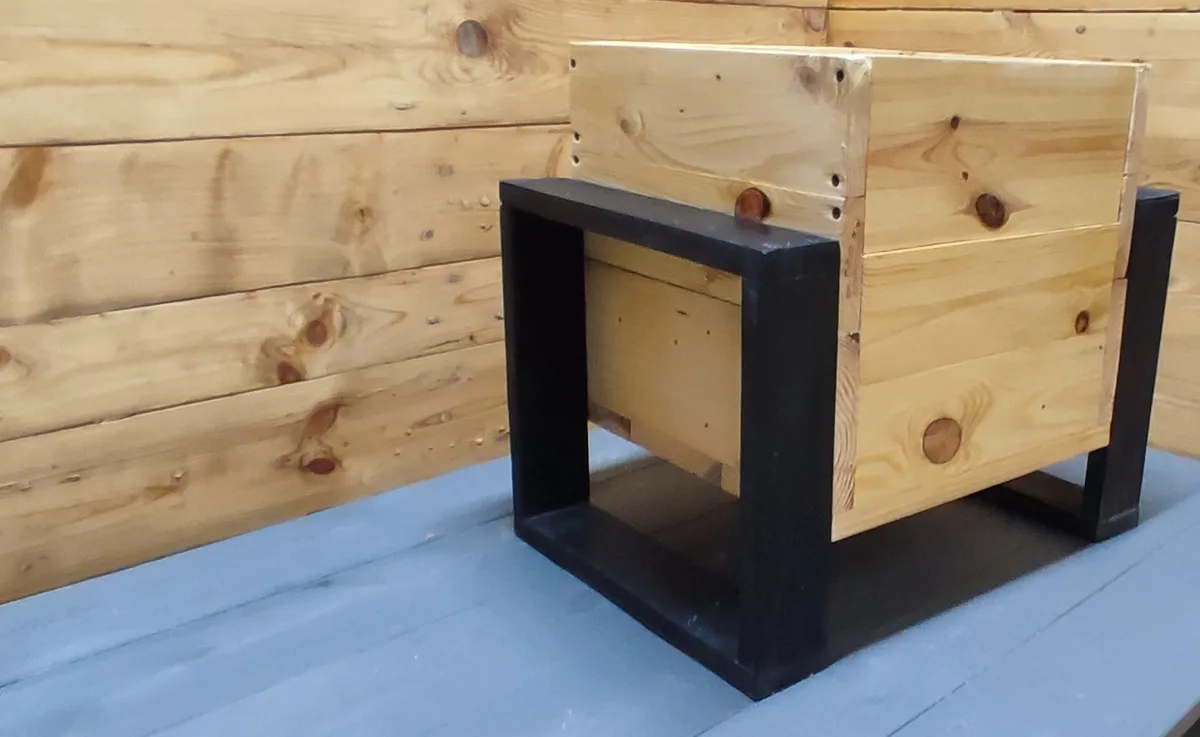 A handmade wooden box with black-painted legs sits on a blue wooden surface against a light wood-paneled wall. The box…
