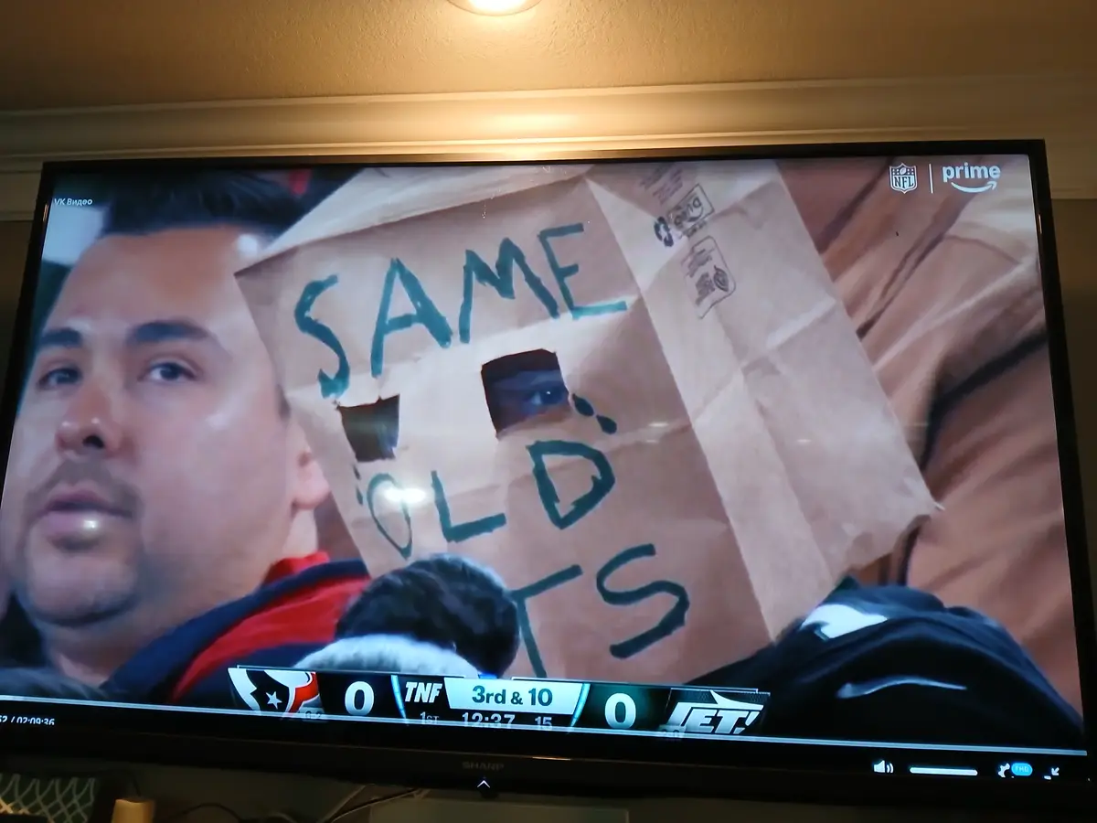 A man is shown on a TV screen holding a cardboard sign with the words 'SAME OLD TS' written in green marker. The sign has…