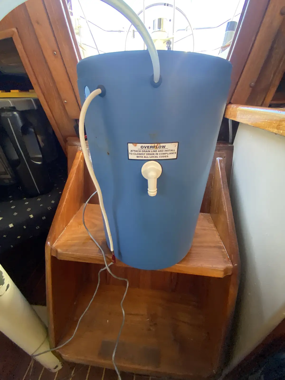 A blue plastic bucket sits on a wooden shelf inside a boat, with a white hose attached to its top and another hose dangling…