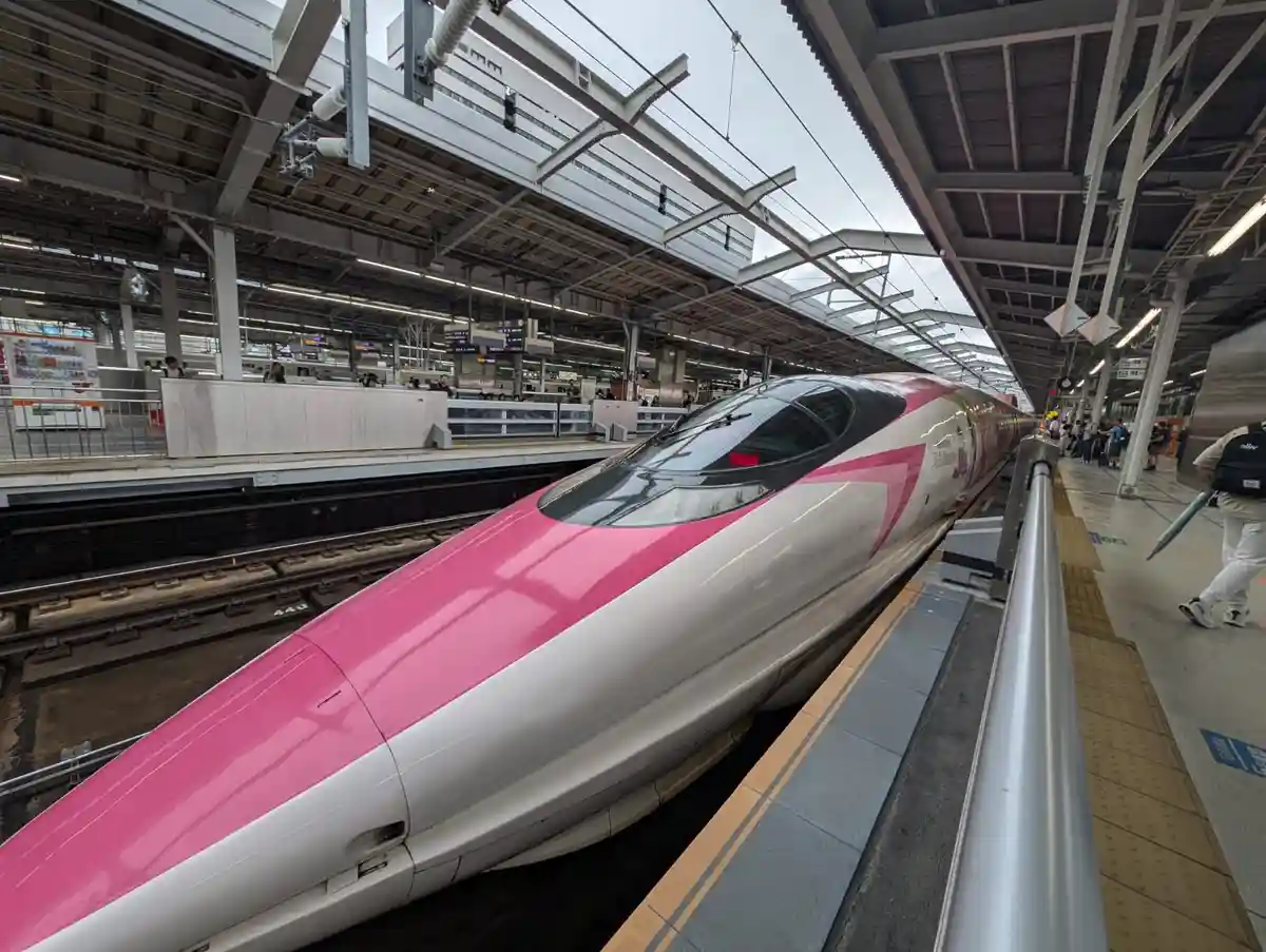 A pink and white high-speed train, likely a Japanese Shinkansen, is stopped at a station platform with passengers nearby…