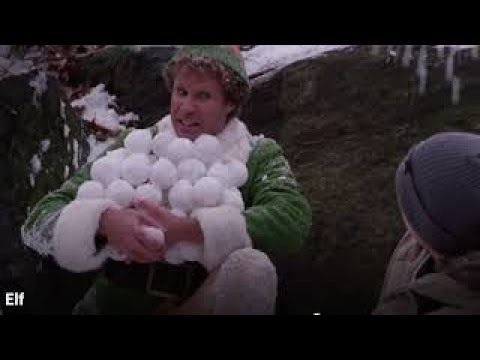 A man in a green elf costume with a white furry collar and hat holds a large pile of snowballs, smiling excitedly while…