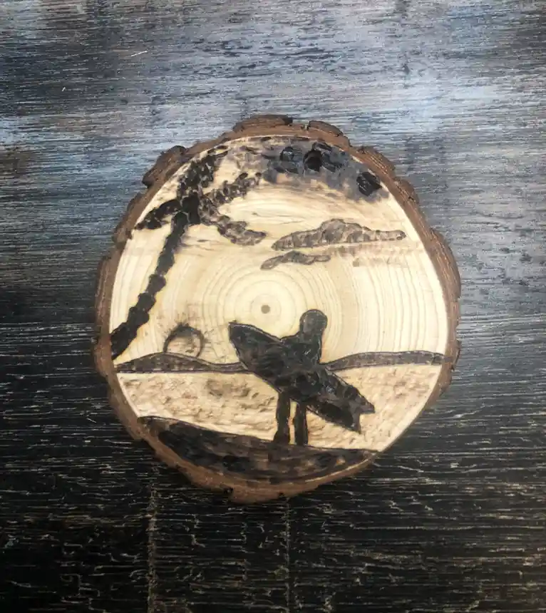 A circular wooden slice with a burned-in scene of a surfer holding a surfboard on a beach, with a palm tree and distant…