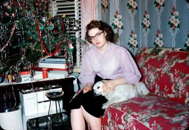 A vintage photo shows a woman with glasses sitting on a red floral couch, holding a small white dog, with a decorated…