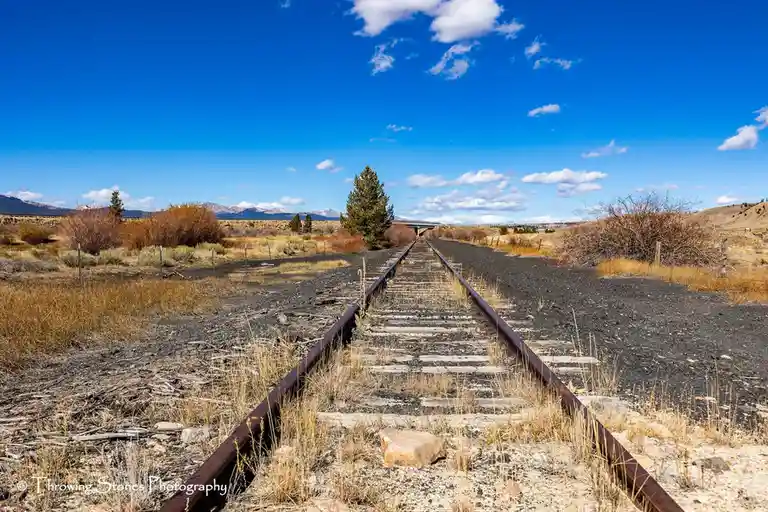An abandoned railway track stretches into the distance through a dry, grassy landscape under a bright blue sky with…