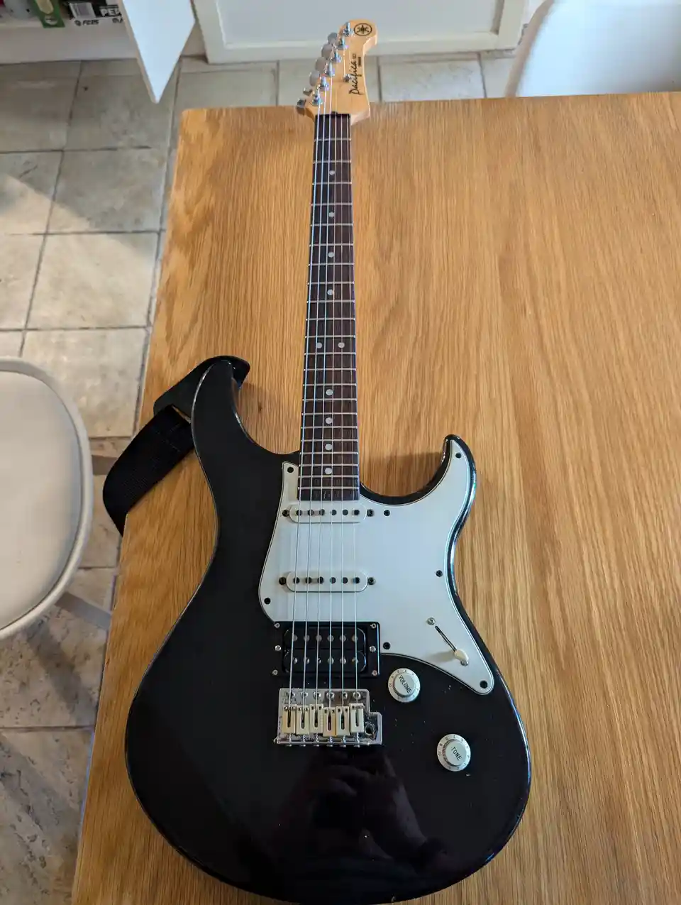 A black Yamaha Pacifica electric guitar with a white pickguard rests on a wooden table, its glossy finish reflecting a…