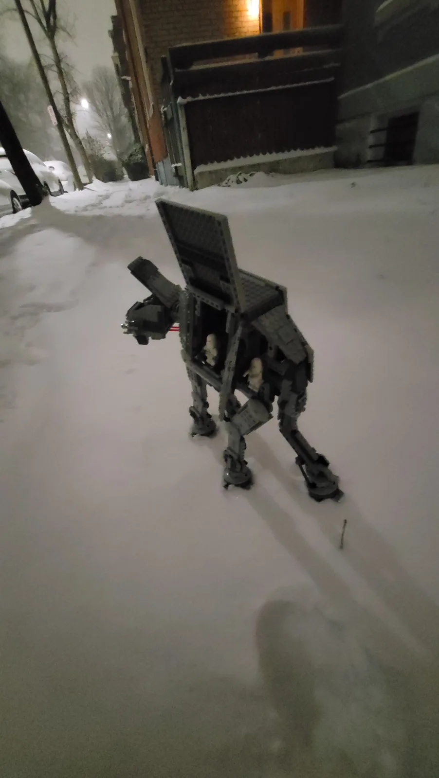 A gray Lego AT-AT walker model stands in deep snow on a residential street at night, with houses and parked cars visible in…
