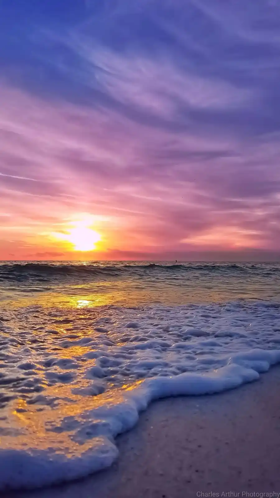 A serene beach sunset photo with vibrant purple, pink, and orange skies over the ocean, waves with white foam rolling onto…