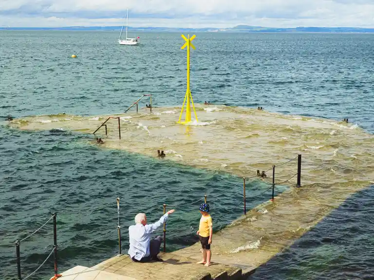 An older man and a young child stand on a concrete pier overlooking choppy water. The man points excitedly toward a bright…