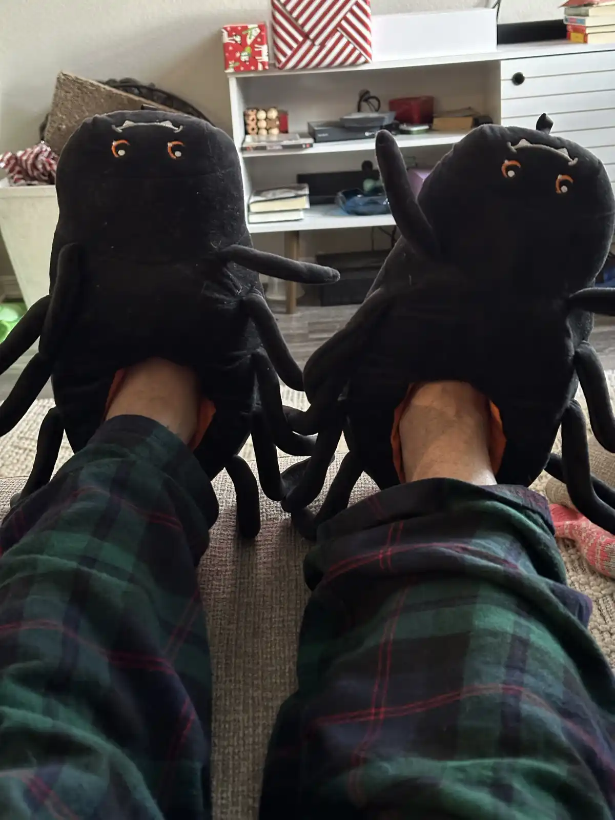 A first-person view of someone wearing oversized black plush spider slippers, with the spider's legs dangling around their…