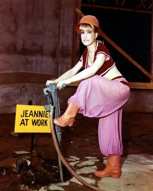 A woman in a retro outfit with a pink skirt, orange hat, and boots is operating a jackhammer in a construction-like setting…