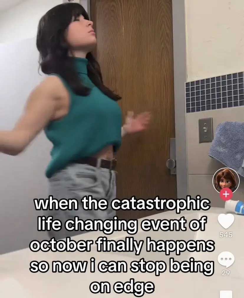 A person in a teal crop top and jeans stands in a bathroom, arms outstretched, head tilted back in a gesture of relief or…