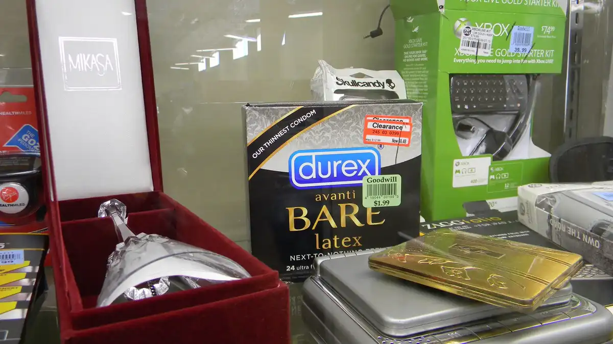 A shelf in a thrift store displays a box of Durex Avanti Bare latex condoms next to a Mikasa crystal trophy, a gold-plated…