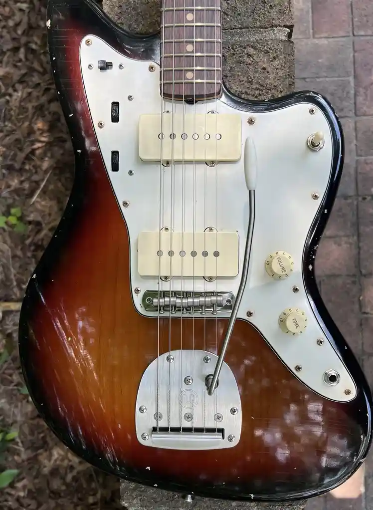 A sunburst-finish electric guitar with a white pickguard and two pickups rests on a brick surface. A long, thin metal rod…