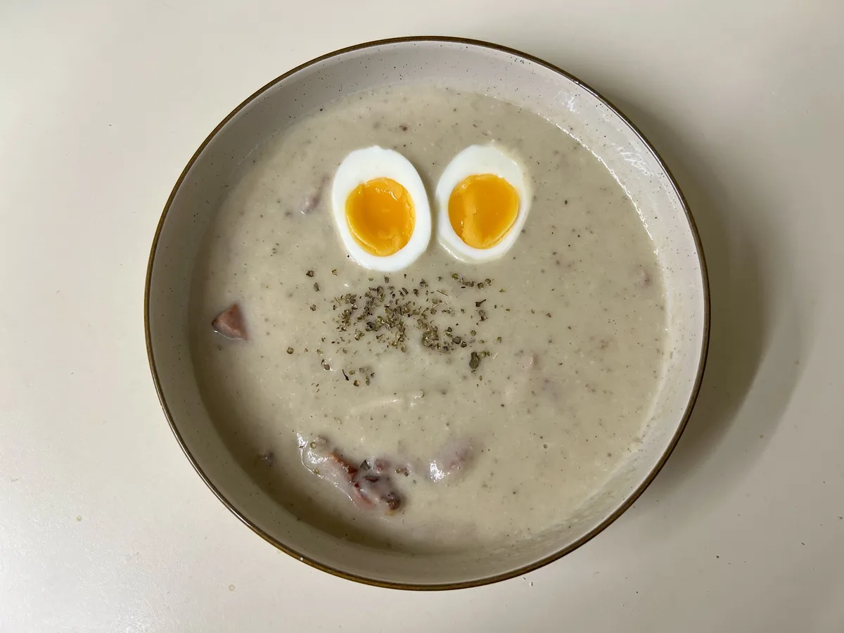 A top-down photo of a creamy soup in a speckled bowl, with two halves of a hard-boiled egg placed as eyes and a sprinkle of…