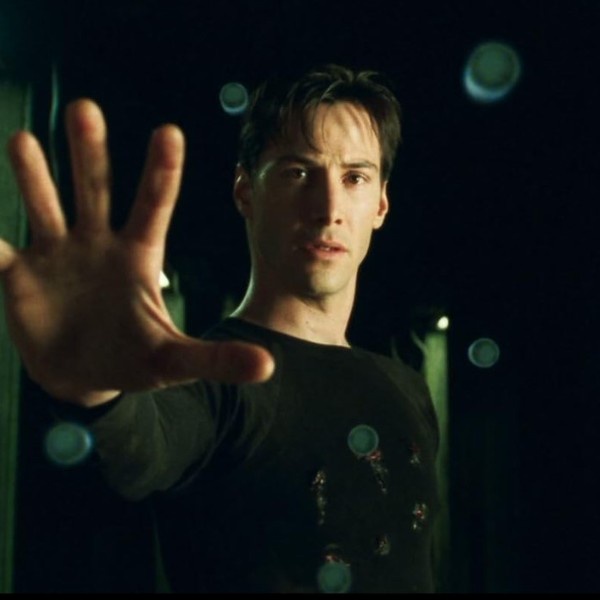 A man with short dark hair and a black t-shirt holds his right hand out flat, palm forward, as if stopping something. His…