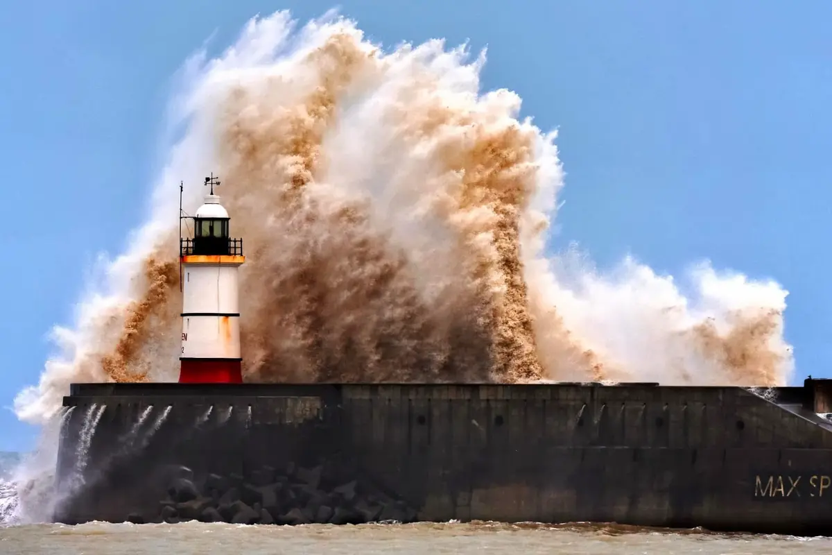 A powerful wave crashes against a concrete sea wall, sending a massive plume of brown water and spray high into the air. A…