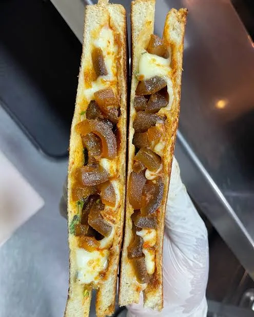 A close-up photo shows a sandwich cut in half, held by a gloved hand, revealing melted cheese and caramelized onions inside…