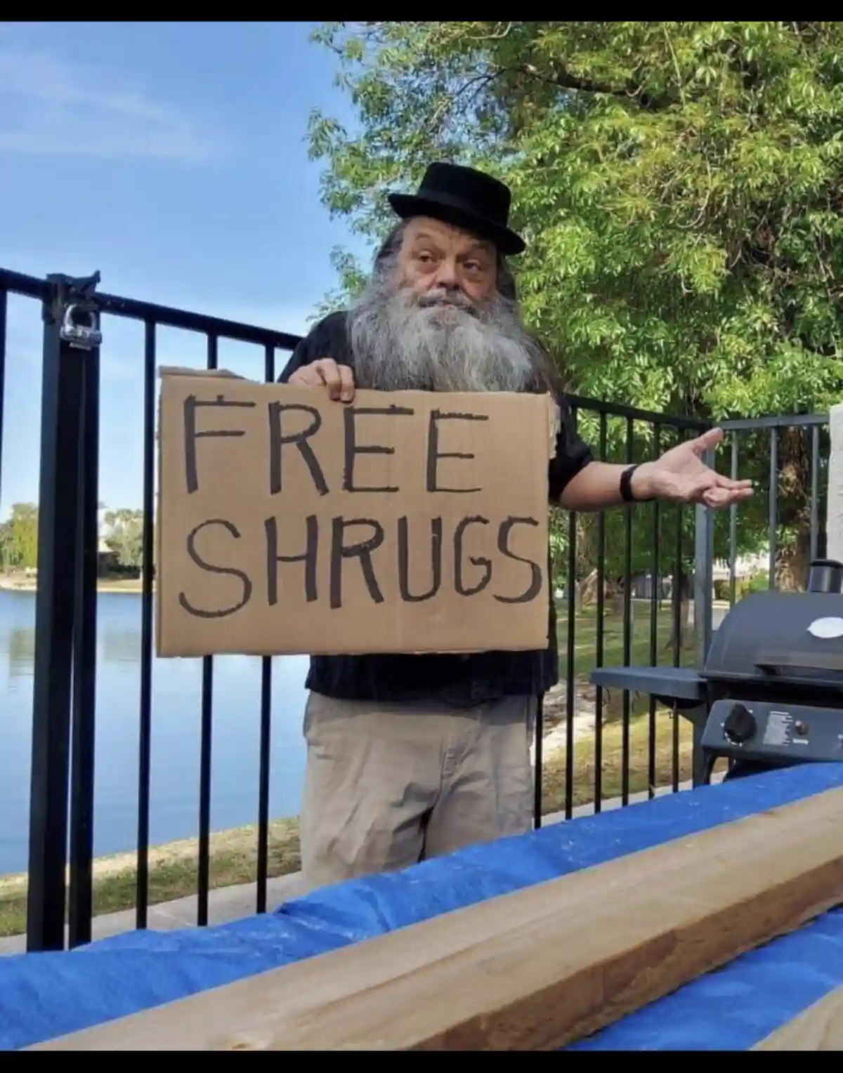 A man with a long gray beard and a black top hat stands outdoors holding a cardboard sign that reads 'FREE SHRUGS'. He…