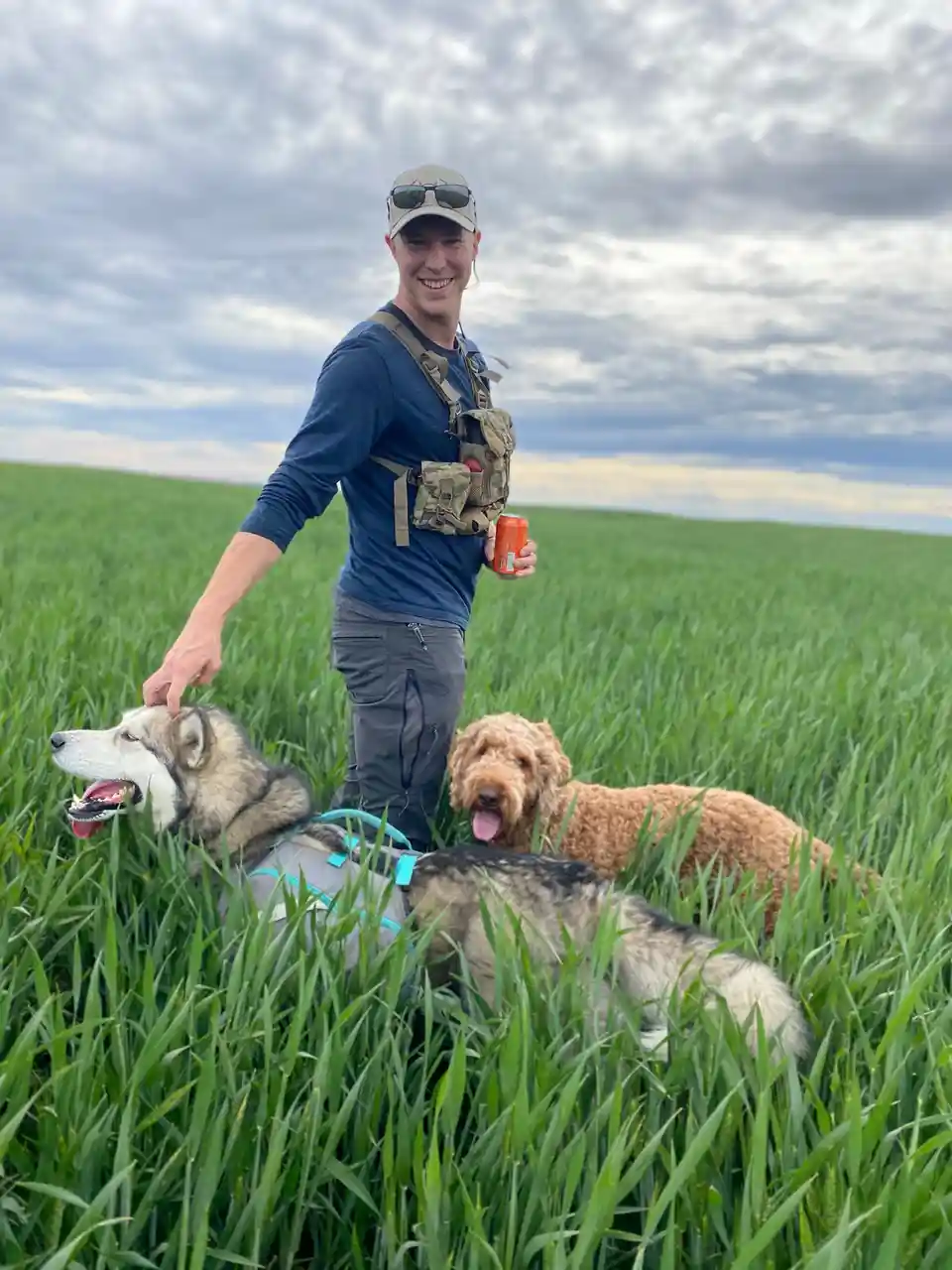 A smiling man in a cap and vest stands in a tall green field, holding an orange can and petting a husky wearing a harness. A…