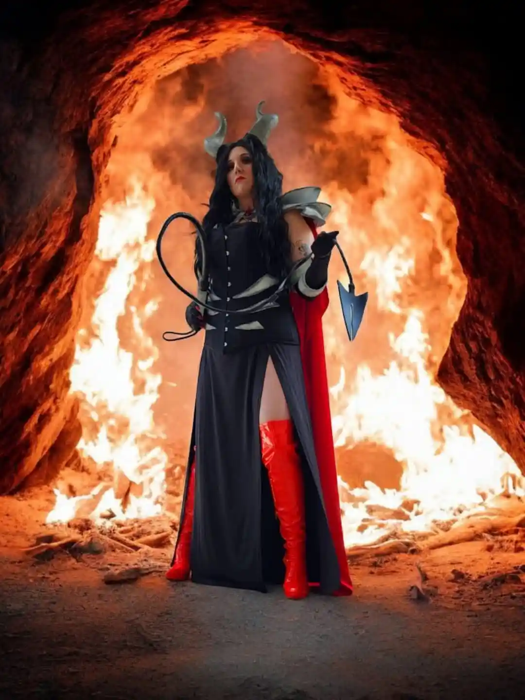 A person in a devil costume with horns, holding a whip and a pitchfork, stands confidently in front of a fiery cave…