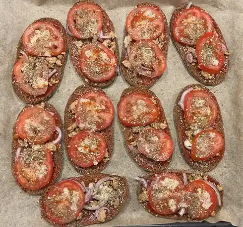 An overhead photo shows nine slices of dark bread arranged on parchment paper, each topped with tomato slices, red onion…