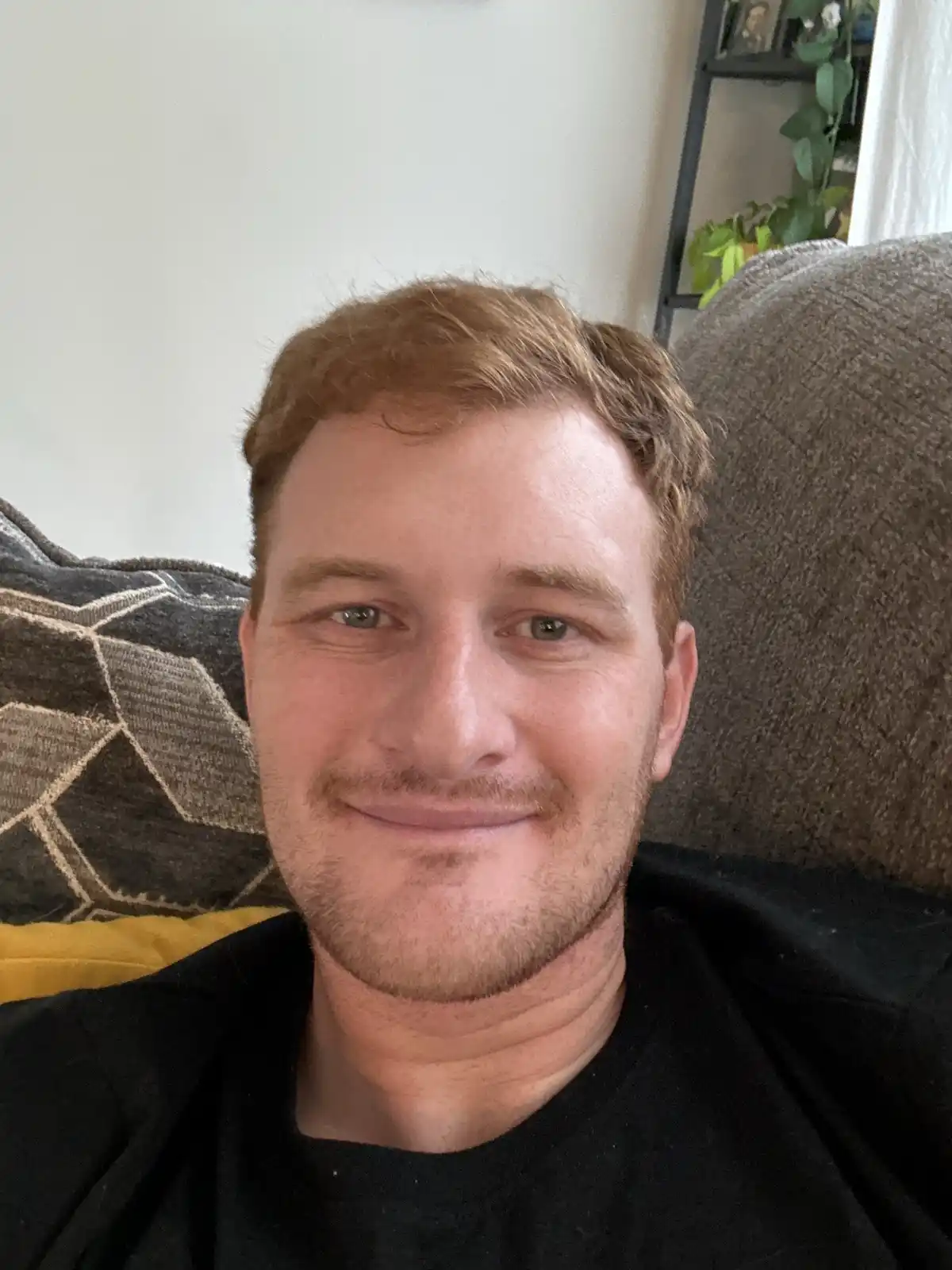 A man with reddish-brown hair and a light beard smiles faintly while sitting on a couch with patterned and solid-colored…