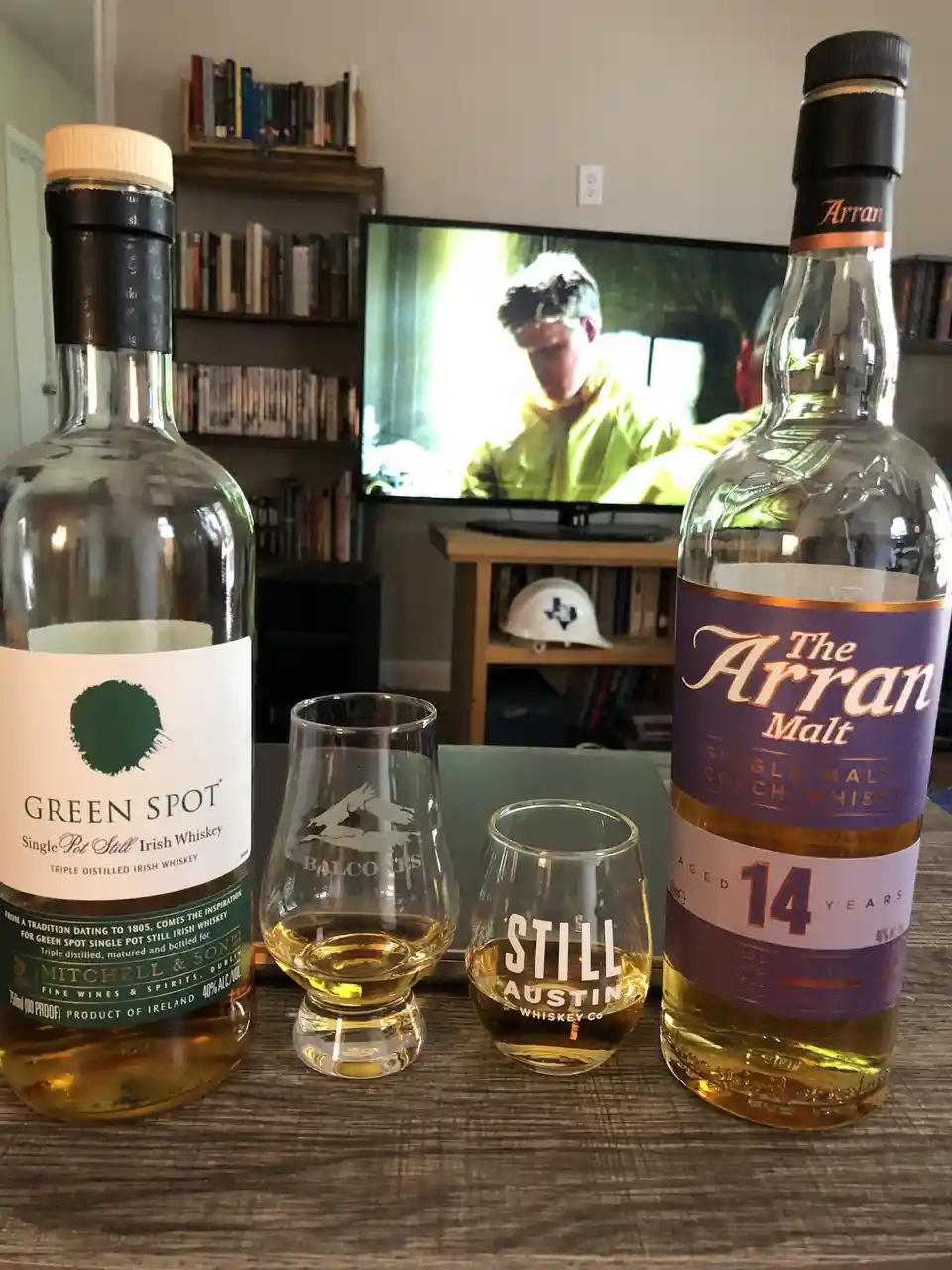 Two whiskey bottles, Green Spot Irish Whiskey and The Arran Malt Scotch Whisky, sit on a wooden table with two whiskey…