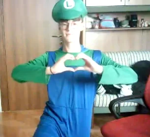 A person dressed as Luigi from Mario games, wearing a green cap with an 'L', green shirt, and blue overalls, forms a heart…