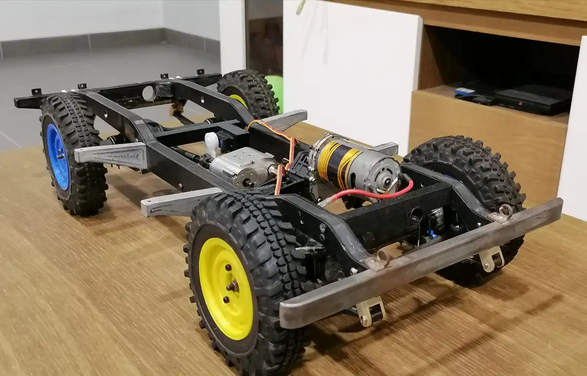 a custom-built remote-controlled car chassis sits on a wooden table, showing exposed motors, wiring, and mismatched…