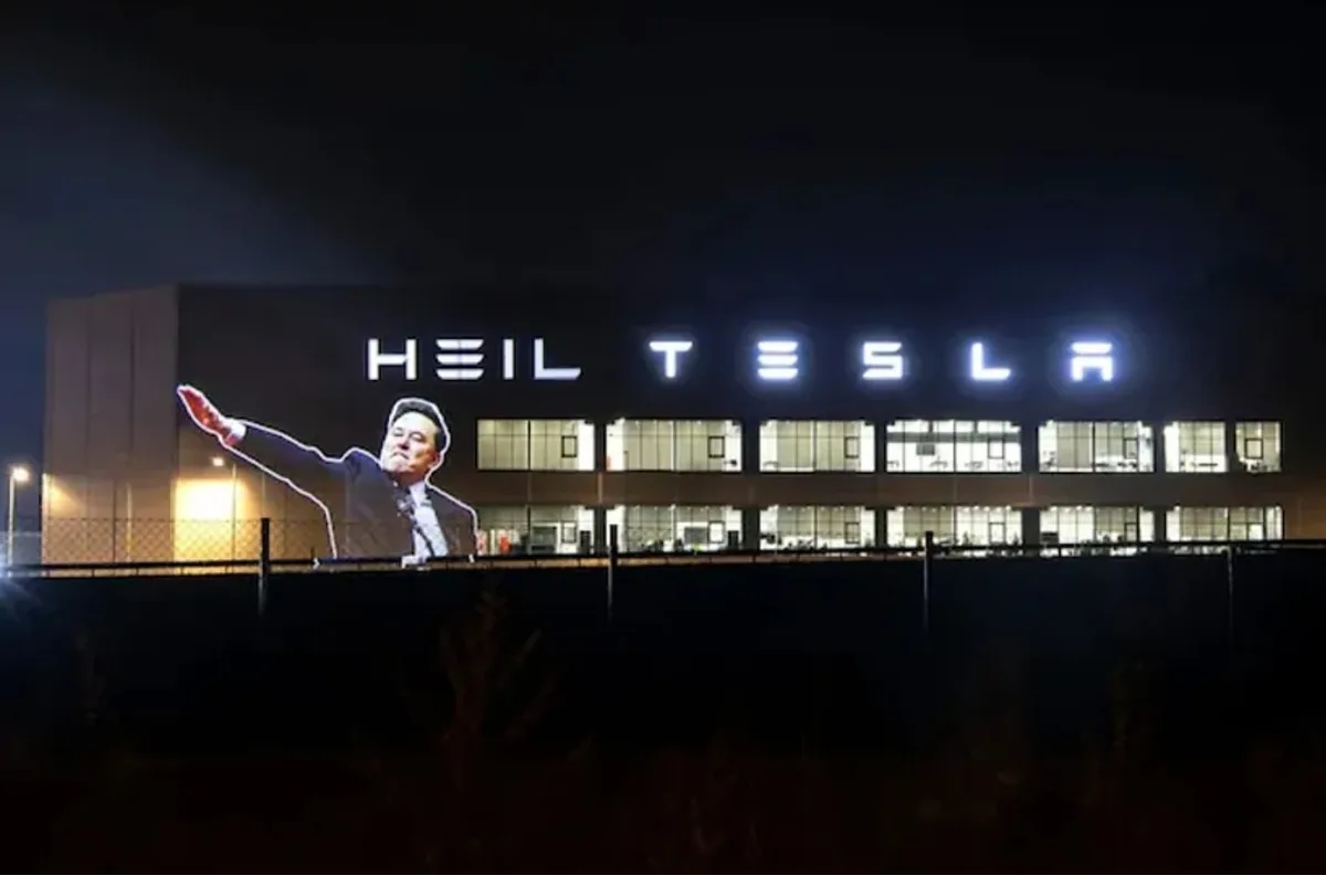A nighttime photo of a large building with illuminated signage reading 'HEIL TESLA'. Superimposed on the building is a…