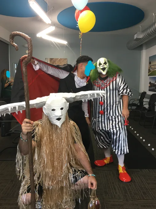 Three people in costumes stand in an office: one as a grim reaper with a scythe and red cape, one as a clown in striped…