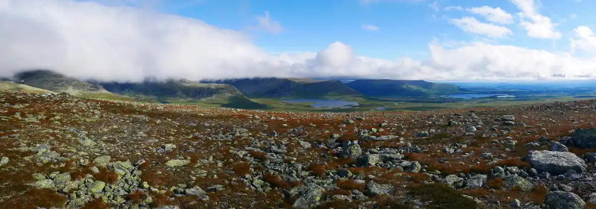 A wide, scenic panorama of a rocky, moss-covered mountain ridge under a partly cloudy blue sky, with distant lakes and…