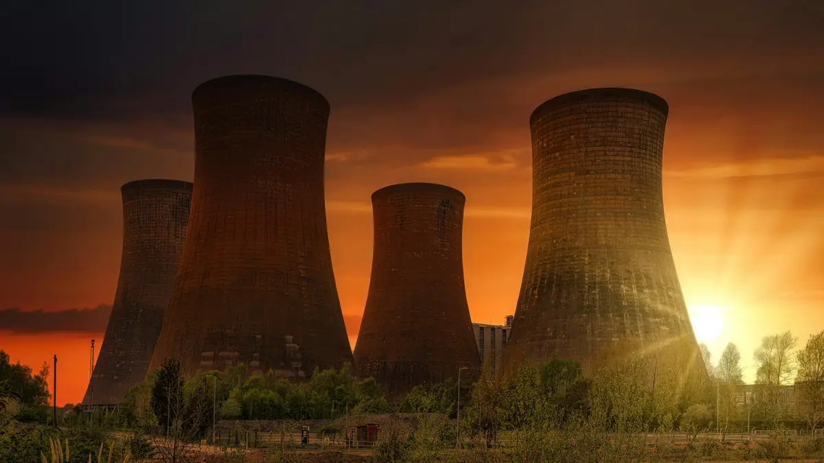 The image shows four large, weathered nuclear power plant cooling towers silhouetted against a dramatic orange and dark sky…