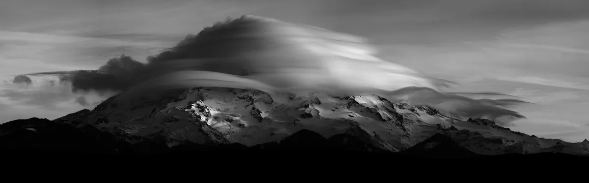 A black-and-white panoramic photograph shows a snow-capped mountain range with dramatic, streaked clouds flowing over its…