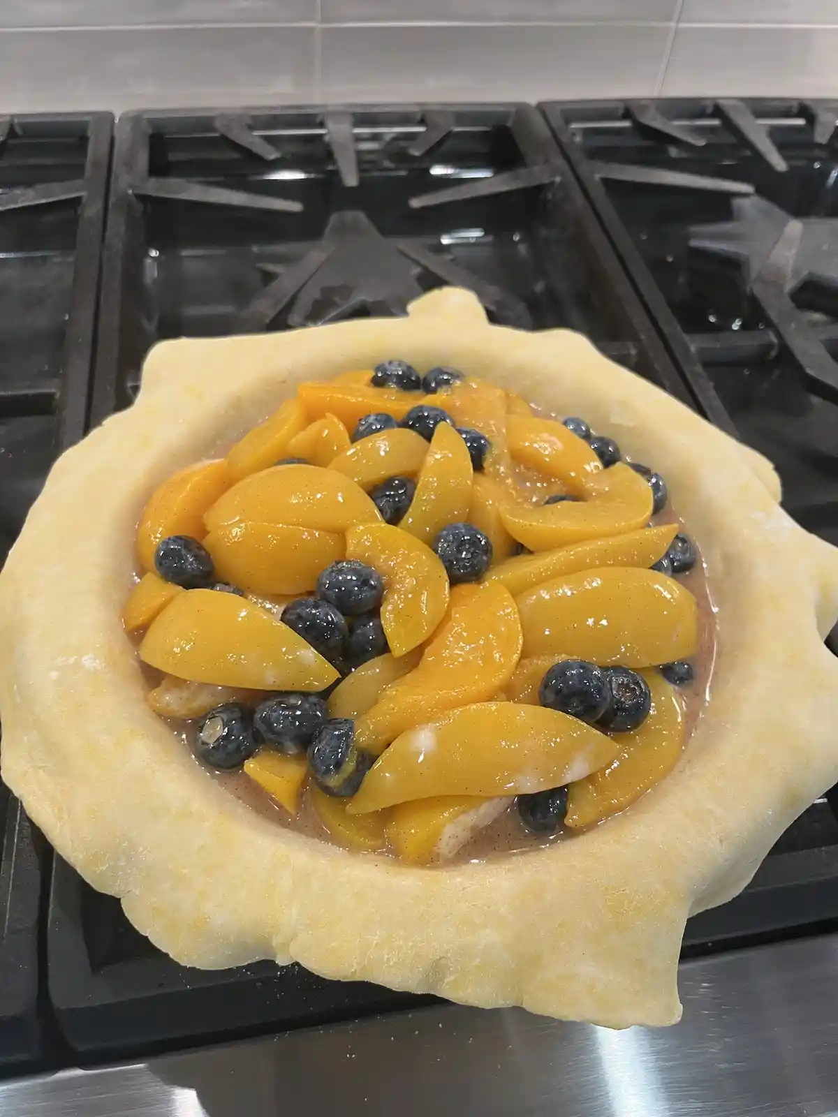 A close-up photo shows an unbaked pie crust on a stovetop, filled with sliced yellow peaches and scattered blueberries. The…