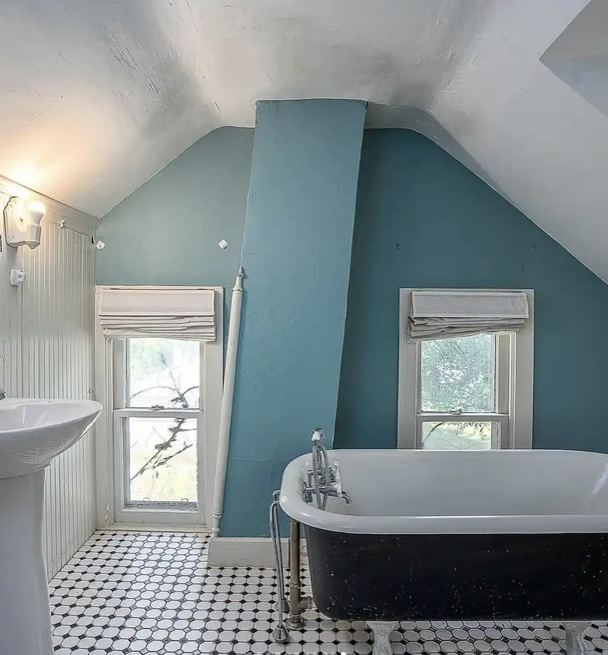 A bathroom with a black clawfoot bathtub, white pedestal sink, and black-and-white hexagonal tile floor, set against teal…