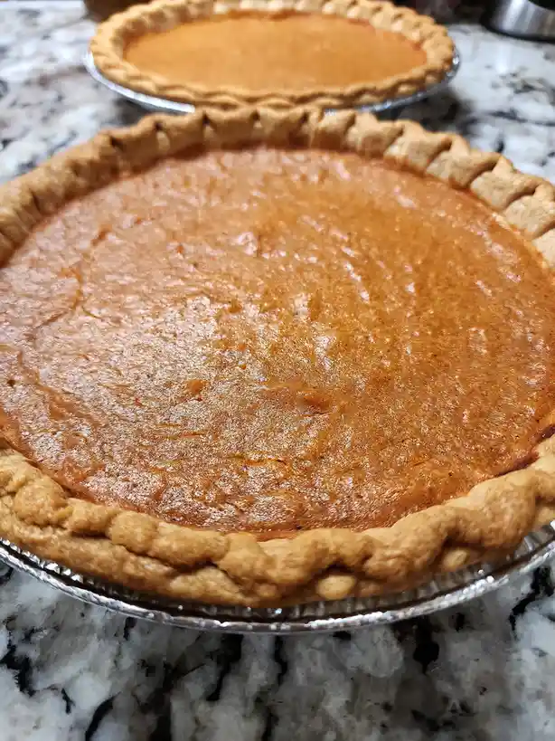 Two freshly baked pumpkin pies sit in aluminum pie tins on a marble countertop, with the foreground pie in sharp focus and a…