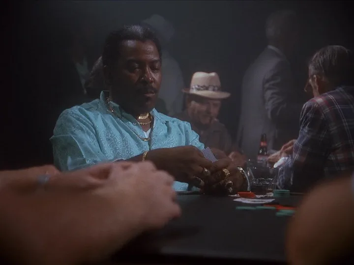 A man in a light blue shirt and gold jewelry sits at a poker table, holding cards with a serious expression. Other players…