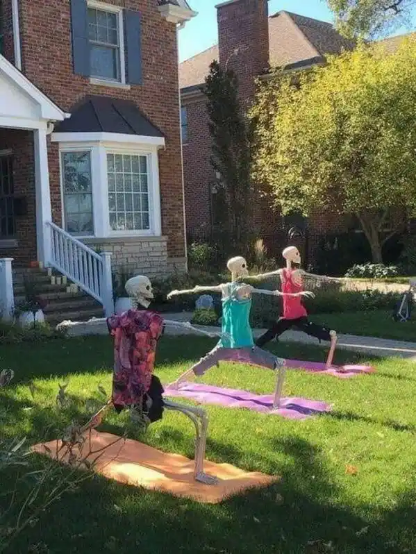 Three skeleton decorations dressed in colorful workout clothes are positioned on yoga mats in a grassy front yard, holding…
