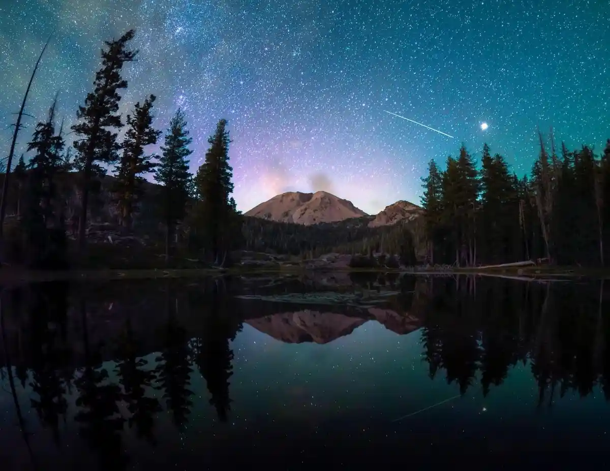 A serene night landscape featuring a star-filled sky with a visible shooting star, reflected perfectly in a calm lake…