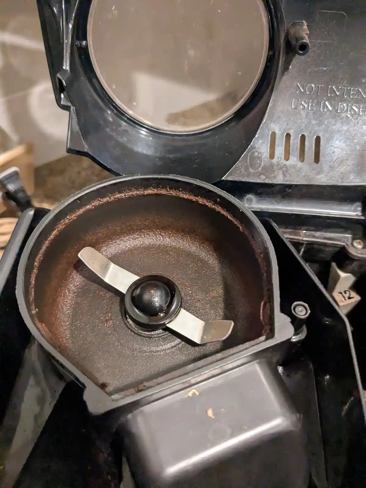 A close-up of a black food processor with its lid open, revealing a bowl coated in a thick, dark brown substance resembling…