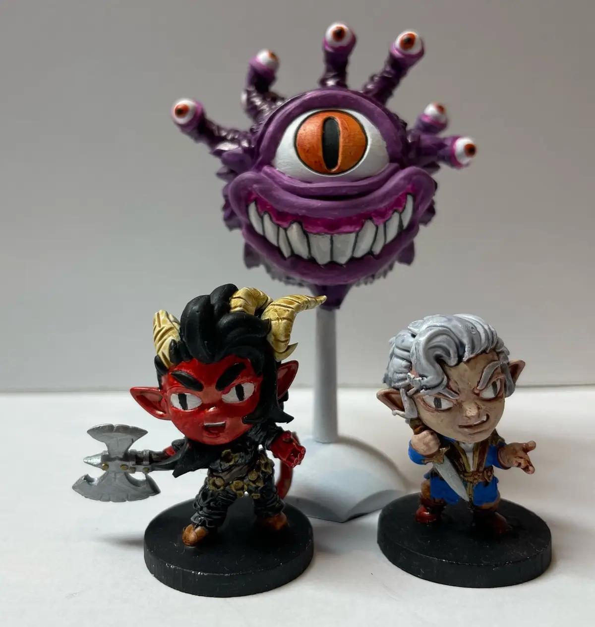 Three chibi-style fantasy miniatures are posed on a white surface: a purple, multi-eyed monster with a giant central eye and…