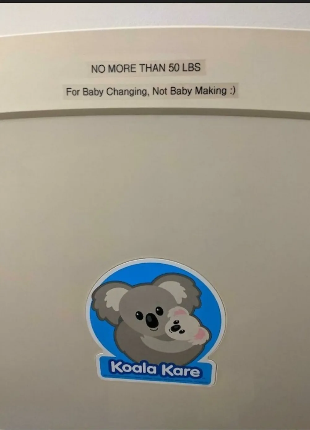 A close-up of a beige baby changing table with a weight limit warning sticker reading 'NO MORE THAN 50 LBS' and a secondary…