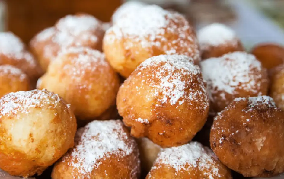 A close-up photo shows a pile of golden-brown, round fried dough balls dusted with powdered sugar. Overlaid text reads…